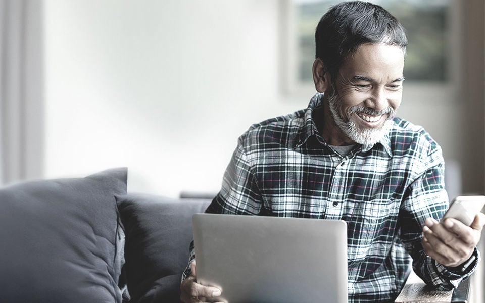Un hombre sonriendo mientras usa el ordenador portátil y el teléfono en casa